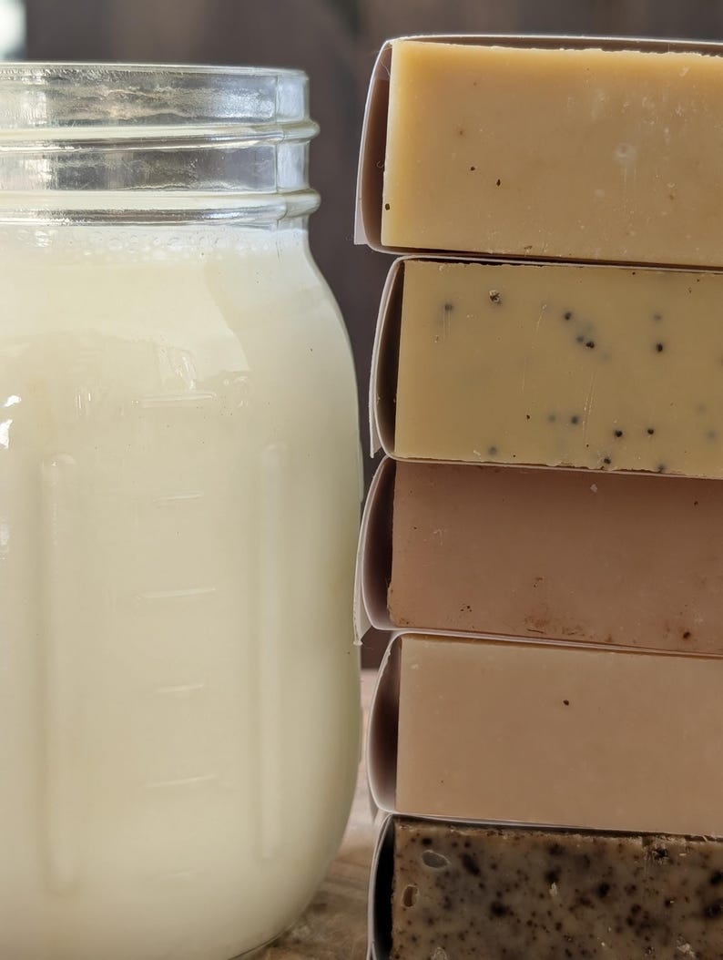 May include: A mason jar filled with a creamy liquid sits next to a stack of wrapped soap bars. The soaps vary in color, from light beige to dark brown, with some containing visible seeds or particles. The jar has measurement markings.