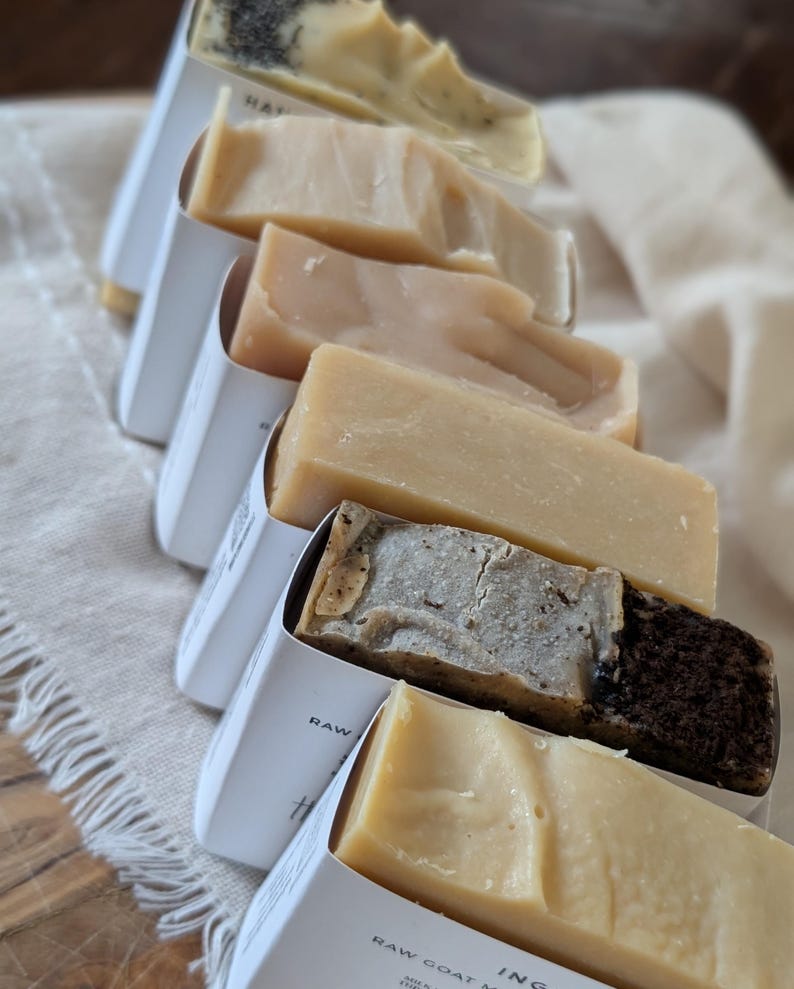May include: Assortment of handmade soaps in white paper packaging. The soaps vary in color, from light beige to dark brown, and are arranged on a neutral-colored cloth. The packaging has text that is partially visible.