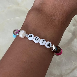 May include: A beaded bracelet with the word "PUTA" spelled out in white letter beads. The bracelet features a mix of colorful beads, including pink, blue, and clear, and a silver star charm.