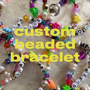 May include: A close-up of several colorful beaded bracelets with various words and charms. The bracelets feature white pearls, colorful beads, and letter beads spelling out words like "QUEER", "SLAY", "PRIDE", and "LOVE".