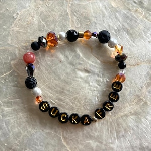 Op de afbeelding: Een kralenarmband met het woord "SUCKAFREE" in gouden letters op zwarte kralen. De armband heeft een verscheidenheid aan kralen in zwart, oranje, wit en roze, met enkele facetgeslepen en getextureerde kralen.
