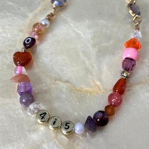 Może przedstawiać: Kolorowa bransoletka z koralików z różnymi koralikami w odcieniach różu, fioletu, pomarańczy i złota. Bransoletka zawiera koralik w kształcie serca, koralik z okiem i koraliki z numerami "415".