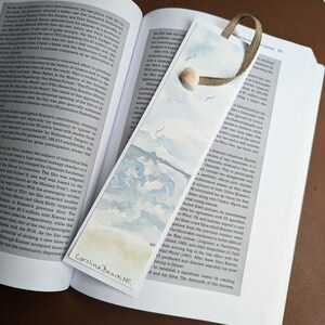 May include: A watercolor bookmark featuring a seascape with a light blue sky, white clouds, and a distant shoreline. The bookmark is decorated with the text "Carolina Beach, NC".