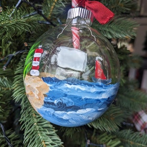 May include: A clear glass Christmas ornament with a painted scene of a lighthouse on a beach with blue waves. A candy cane is inside the ornament.