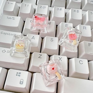 Może przedstawiać: Cztery przezroczyste klawisze z różnymi różowymi i białymi wzorami kreskówek na białej klawiaturze. Wzory obejmują motyla, lizak, ciasto i lizak w kształcie serca.