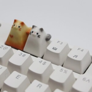 May include: Two small ceramic cat figurines, one orange and white, the other white and gray, are placed on top of a white keyboard. The orange and white cat is on the "3#" key, and the white and gray cat is on the "4$" key.