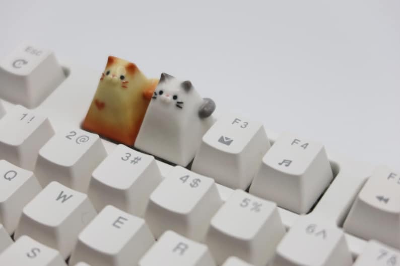 May include: Two small ceramic cat figurines, one orange and white, the other white and gray, are placed on top of a white keyboard. The orange and white cat is on the "3#" key, and the white and gray cat is on the "4$" key.