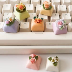 以下が含まれることがあります： 花柄のカラフルなキーキャップが6つ。キーキャップは白いキーボードの上に並んでいます。キーキャップはプラスチック製で光沢のある仕上げになっています。花は布製で、リアルな外観をしています。