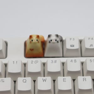 May include: Two small ceramic cat keycaps, one orange and white, the other gray and white, are placed on a white keyboard. The keycaps are on the keys labeled "2@" and "3#" on the keyboard.