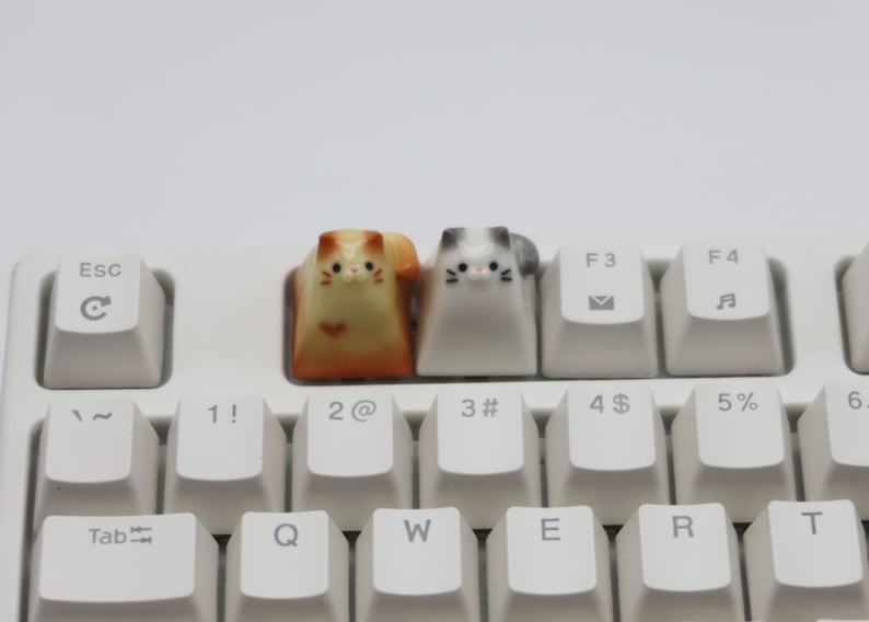 May include: Two small ceramic cat keycaps, one orange and white, the other gray and white, are placed on a white keyboard. The keycaps are on the keys labeled "2@" and "3#" on the keyboard.
