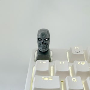 May include: A grey, skull-shaped keycap sits on a white keyboard. The keycap has glowing white eyes and a detailed, metallic appearance. The keyboard keys include Tab, 1!, 2@, and F1.