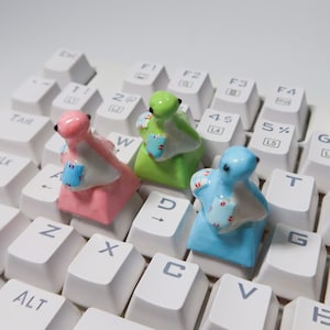 May include: Three small ceramic keycaps shaped like animals, each with a different colour and pattern. The keycaps are sitting on a white keyboard.