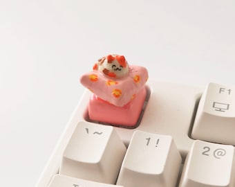 May include: A small, pink, and orange cat figurine is sleeping on a pink pillow. The figurine is sitting on a white keyboard.