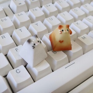 May include: Two small ceramic cat keycaps in white and orange with black eyes and whiskers. The keycaps are on a white keyboard.