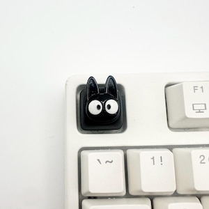May include: A black custom keycap shaped like a cat head with large white eyes and pointed ears, installed on a white mechanical keyboard next to a key labeled F1 and a key labeled with the number 1 and an exclamation point symbol.