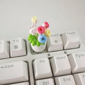 Puede incluir: Un adorno de Pascua caprichoso con una pequeña maceta blanca con tulipanes coloridos y una figura de conejito. Los tulipanes son rosas, amarillos y azules, con hojas verdes. El adorno está sobre un teclado blanco.