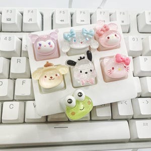 May include: A selection of seven decorative keyboard keycaps with cartoon characters. The keycaps are in a white tray, resting on a white keyboard. The characters include a frog, a dog, a cat, and other cute designs. The keycaps are made of a translucent material.