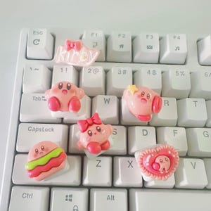 Pode incluir: Uma coleção de teclas de teclado cor-de-rosa com o tema Kirby num teclado branco. As teclas apresentam vários designs, incluindo Kirby com uma estrela, um laço e um hambúrguer. A palavra "Kirby" também é exibida.
