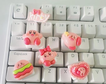 May include: A collection of pink Kirby-themed keycaps on a white keyboard. The keycaps feature various designs, including Kirby with a star, a bow, and a burger. The word "Kirby" is also displayed on a keycap.