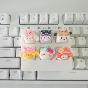 Pode incluir: Um conjunto de seis teclas decorativas e coloridas com personagens de desenhos animados. As teclas estão em um suporte branco, em um teclado branco. Os personagens incluem um coelho, um pinguim, um urso, um cachorro, um gato e um personagem com um chapéu rosa.