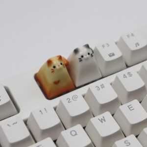May include: Two small cat-shaped keycaps for a keyboard. The keycaps are white and orange with black eyes and whiskers. The keycaps are on a white keyboard with black keys.