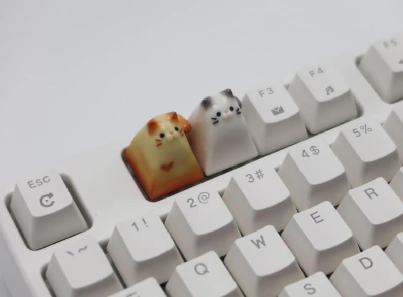 May include: Two small cat-shaped keycaps for a keyboard. The keycaps are white and orange with black eyes and whiskers. The keycaps are on a white keyboard with black keys.