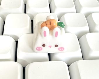 May include: A white bunny keycap with a carrot on top. The keycap is sitting on a white keyboard.