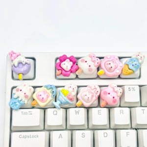 May include: A white keyboard with custom keycaps shaped like colorful ice cream treats. The keycaps feature various designs, including popsicles, ice cream cones, and floral shapes, in shades of pink, blue, and purple.