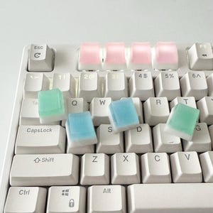 May include: A white keyboard with pastel blue and green keycaps. The keycaps are translucent and have a frosted finish. The keyboard has a standard layout with keys for letters, numbers, and symbols.