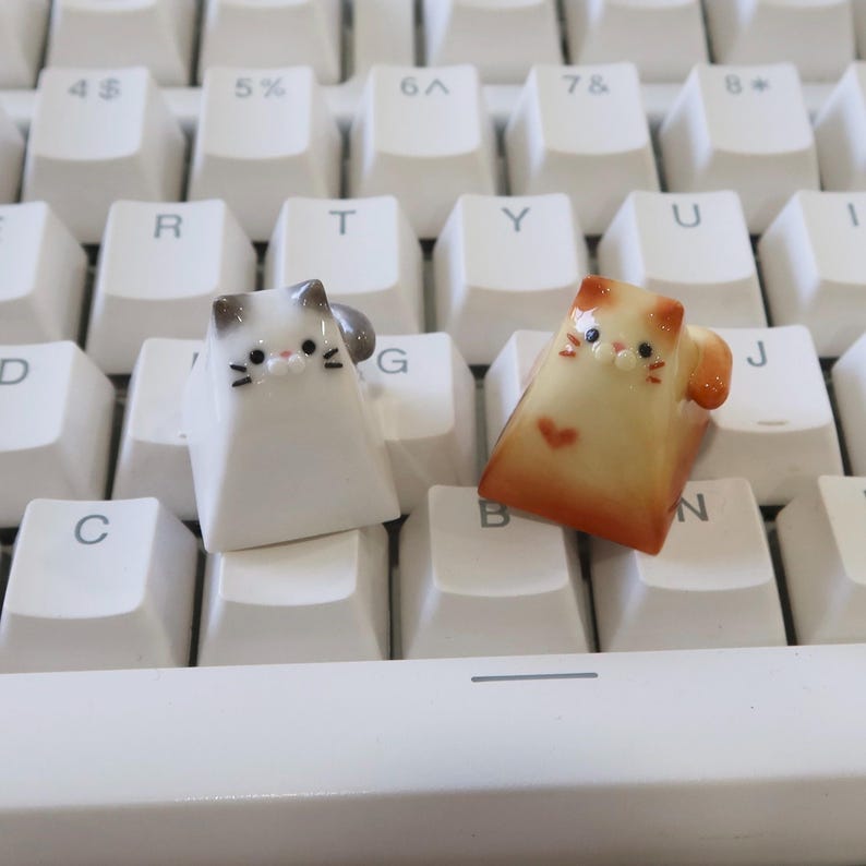 May include: Two ceramic cat keycaps, one white with gray ears and one light orange with a pink heart on its chest, are placed on a white keyboard.