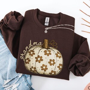 May include: A dark chocolate brown crewneck sweatshirt featuring a graphic of a cream-colored pumpkin decorated with brown daisy flowers and the word grandma written in cursive script. The garment has long sleeves and a ribbed collar and cuffs.