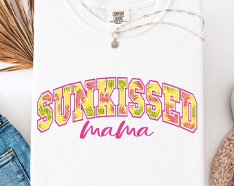 May include: White crew neck sweatshirt with the text "Sunkissed Mama" in a colorful, tropical-themed design. The sweatshirt is folded, with a silver necklace and a pair of white sneakers nearby. A pair of blue jeans and a dried palm leaf are also in the image.