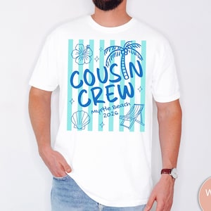Op de afbeelding: Een wit t-shirt met een afbeelding van lichtblauwe verticale strepen en de donkerblauwe tekst COUSIN CREW Myrtle Beach 2026. Het ontwerp bevat illustraties van een hibiscusbloem, een palmboom, een zeeschelp en een strandstoel.