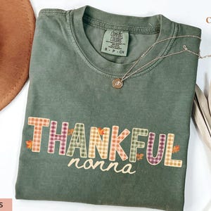 May include: A moss green short-sleeve t-shirt featuring the word THANKFUL in a multi-colored plaid pattern with small orange leaf accents, and the word nonna written in a cursive white font below. The shirt has a crew neckline and a tag.
