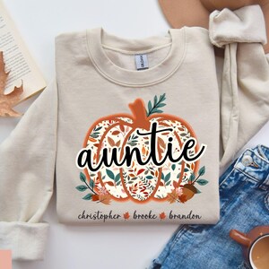 May include: Sand-colored sweatshirt with a pumpkin design. The pumpkin is filled with fall leaves and the word "auntie" in script. Below the pumpkin are the names "Christopher", "Brooke", and "Brandon".