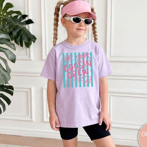 Op de afbeelding: Een lichtpaars t-shirt met een afbeelding van verticale turquoise strepen en de roze tekst "COUSIN CREW" in het midden op de voorzijde. Het ontwerp bevat kleine lijntekeningen van palmbomen, schelpen en bloemen rondom de tekst.