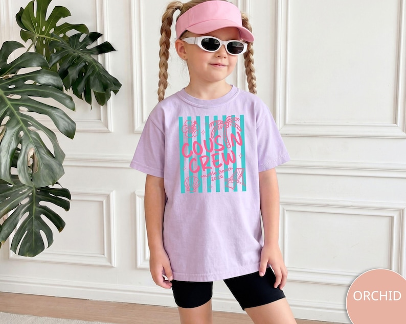 Op de afbeelding: Een lichtpaars t-shirt met een afbeelding van verticale turquoise strepen en de roze tekst "COUSIN CREW" in het midden op de voorzijde. Het ontwerp bevat kleine lijntekeningen van palmbomen, schelpen en bloemen rondom de tekst.
