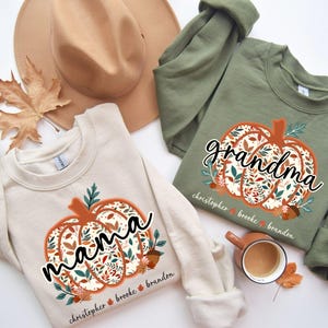 May include: Two crewneck sweatshirts, one cream and one olive green, feature a pumpkin design filled with floral patterns. The cream sweatshirt says "mama" and the green one says "grandma". Both have names below the pumpkin.