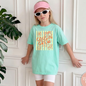 Op de afbeelding: Een licht mintgroen T-shirt met korte mouwen voorzien van een afbeelding met gele verticale strepen, oranje tekst COUSIN CREW en illustraties van een palmboom, hibiscusbloem, schelp en strandstoel met de tekst Myrtle Beach 2025.