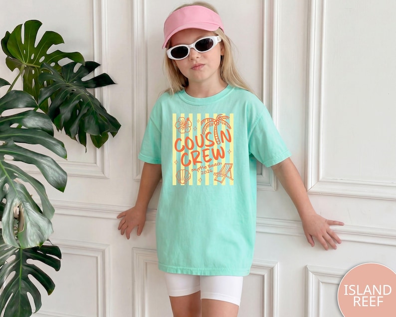 Op de afbeelding: Een licht mintgroen T-shirt met korte mouwen voorzien van een afbeelding met gele verticale strepen, oranje tekst COUSIN CREW en illustraties van een palmboom, hibiscusbloem, schelp en strandstoel met de tekst Myrtle Beach 2025.