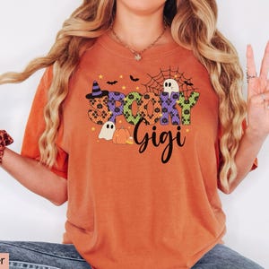Może przedstawiać: Pomarańczowa koszulka z napisem "Spooky Gigi" w halloweenowym wzorze. Wz&oacute;r zawiera kapelusz czarownicy, nietoperze, pajęczynę, ducha i dynię. Koszulka jest w stylu casual.