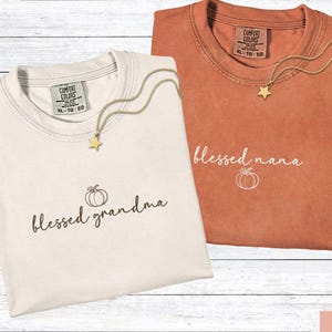 May include: Two folded t-shirts, one ivory and one yam-colored, with the words "blessed grandma" and "blessed nana" printed on them. Each shirt has a pumpkin graphic and a gold star necklace.