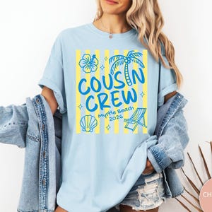 Op de afbeelding: Een lichtblauw t-shirt met een grafische print van gele verticale strepen en blauwe illustraties van een hibiscusbloem, een palmboom, een schelp en een strandstoel. De tekst luidt "COUSIN CREW" en "Myrtle Beach 2026".