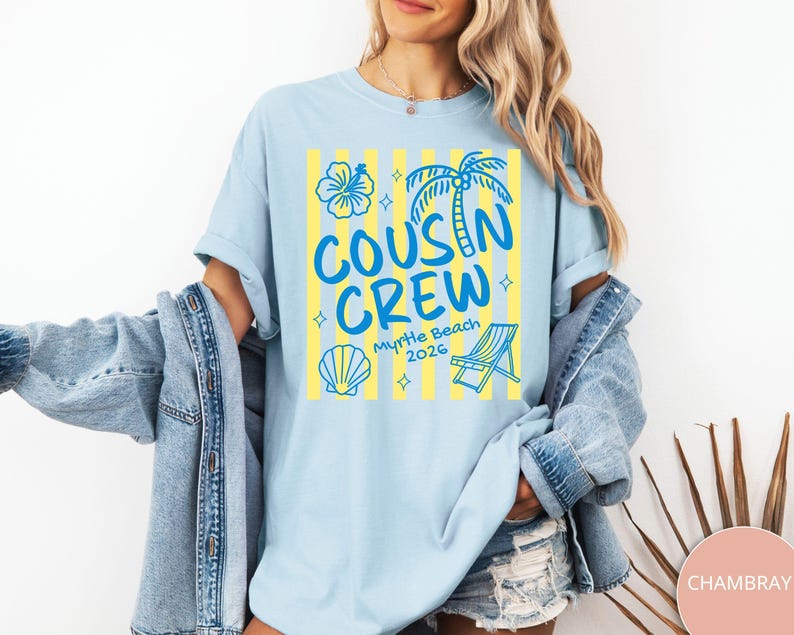 Op de afbeelding: Een lichtblauw t-shirt met een grafische print van gele verticale strepen en blauwe illustraties van een hibiscusbloem, een palmboom, een schelp en een strandstoel. De tekst luidt "COUSIN CREW" en "Myrtle Beach 2026".