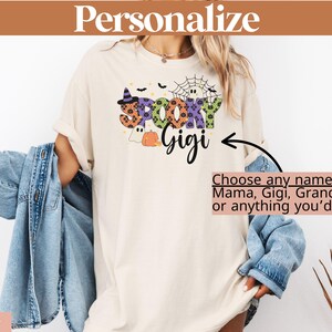 Może przedstawiać: Kremowa koszulka z halloweenową grafiką z napisem "Spooky Gigi". Projekt zawiera kapelusz czarownicy, pajęczynę, ducha i dynię. Tekst "Personalize" znajduje się na g&oacute;rze, a "Choose any name" znajduje się pod grafiką.