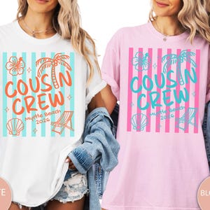 Op de afbeelding: Een wit t-shirt en een lichtroze t-shirt, elk met een grafische print van verticale strepen, een hibiscusbloem, een palmboom, een schelp en een strandstoel. De tekst luidt COUSIN CREW Myrtle Beach 2026 in oranje of blauwgroene inkt.