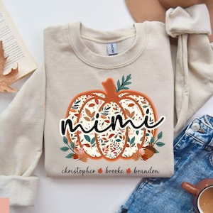 May include: Sand-colored sweatshirt with a pumpkin design. The pumpkin is filled with a floral pattern and the word "mimi" in black script. The names "christopher brooke brandon" are below the pumpkin.