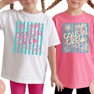 Op de afbeelding: Een wit t-shirt en een roze t-shirt, elk met een afbeelding van verticale turquoise strepen, een hibiscusbloem, een palmboom, een schelp en een strandstoel. De tekst luidt COUSIN CREW Myrtle Beach 2026 in het roze en in het geel.