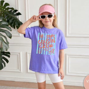 Op de afbeelding: Een violet t-shirt met een grafische print met de tekst COUSIN CREW in oranje, vergezeld van lichtblauwe verticale strepen en oranje illustraties van een palmboom, een strandstoel en een bloem, gecombineerd met een witte korte broek en een roze vizier.