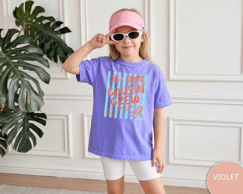 Op de afbeelding: Een violet t-shirt met een grafische print met de tekst COUSIN CREW in oranje, vergezeld van lichtblauwe verticale strepen en oranje illustraties van een palmboom, een strandstoel en een bloem, gecombineerd met een witte korte broek en een roze vizier.